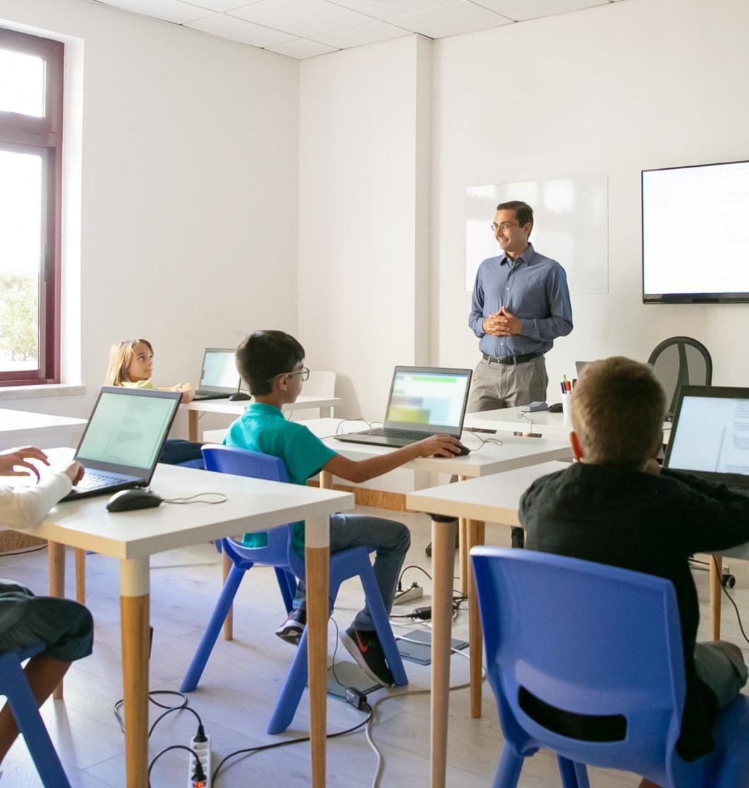 Confident teacher explaining lesson to pupils. Multiethnic children sitting at table in classroom, listening middle-aged man and using laptop computers. Childhood and digital education concept